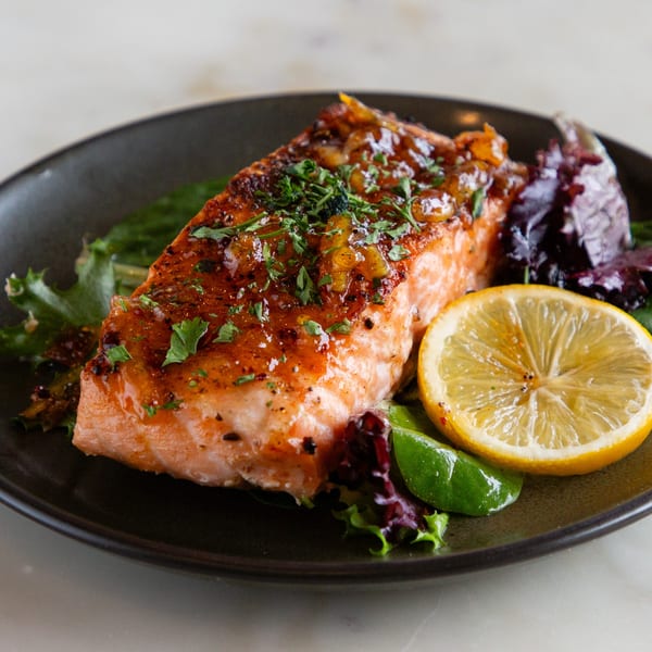 Glazed salmon fillet with herbs, a lemon slice, and mixed greens on a dark plate.