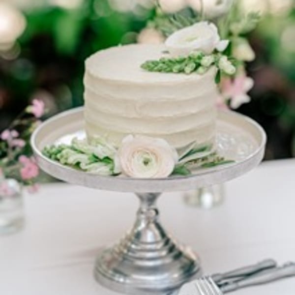 Small white frosted cake on a silver stand, decorated with white flowers and green foliage.